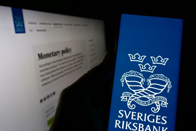 Person håller i smartphone med Sveriges Riksbanks logga på skärmen. I bakgrunden syns Riksbankens hemsida.